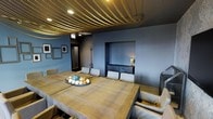 This dining room makes an immediate impression with its eye-catching ceiling treatment, where rows of thick golden rope are stretched across the overhead surface in parallel lines, lending the space a nautical yet refined character. The walls are finished in a deep blue-gray tone that establishes a moody, intimate ambiance throughout. At the center of the room sits a large, solid wood dining table with a beautifully weathered, rustic finish, surrounded by upholstered chairs in neutral tones that provide a soft contrast to the room's darker palette. A decorative bowl holding spherical ornaments serves as a tasteful centerpiece. Along one wall, a recessed niche houses a dark credenza accented by a small table lamp, adding a warm glow and serving as both functional storage and a design focal point. Framed artwork adorns the blue-gray walls, contributing to the curated feel of the space. Recessed lighting integrated into the rope ceiling provides functional illumination. The overall design blends industrial and nautical influences with warm, natural materials to create a dining environment that is both dramatic and welcoming.