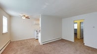 This single-floor space presents an open, functional layout designed for comfortable everyday living. The living area is finished with wall-to-wall carpet in a neutral beige tone, creating a warm and cohesive feel throughout. A ceiling fan with an integrated light fixture is centrally positioned, providing both air circulation and ambient lighting to the main living space.

A partial dividing wall separates the living area from the kitchen without fully closing off the space, maintaining an open and airy atmosphere. The kitchen features white cabinetry and a white electric range, offering a clean, bright aesthetic. The layout allows for easy movement between the two areas.

Baseboard heating units run along the lower portion of the walls, ensuring consistent warmth throughout the space. The walls are painted in a crisp, neutral white, complementing the light-toned flooring and cabinetry. A doorway visible toward the back of the space suggests access to an additional room or area. Overall, the space maintains a clean, straightforward design with practical features suited for daily living.