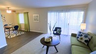 This single-floor space offers a thoughtfully designed interior that balances comfort and functionality. The living area serves as the heart of the home, featuring a generous open layout that creates an airy, spacious feel. Large sliding glass doors dressed with vertical blinds allow abundant natural light to pour in, brightening the room and offering a view of the outdoors. The flooring transitions seamlessly between plush carpet in the main seating area and smooth hardwood-style flooring, adding visual interest and practical versatility to the space. Neutral wall tones provide a clean, adaptable backdrop that complements a range of décor styles. A second window dressed with vibrant green curtains adds a pop of color and draws in additional natural light from another angle. The layout accommodates both relaxed lounging and easy movement throughout. Overall, the space presents a warm and inviting ambiance, well-suited for everyday living, with its emphasis on natural light, open flow, and a calm, contemporary aesthetic that makes the interior feel both functional and welcoming.