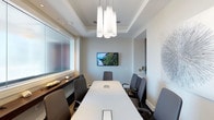 This polished conference room offers a refined and professional environment ideal for focused meetings and collaborative sessions. The space is anchored by a long, white rectangular conference table with built-in connectivity ports, surrounded by eight cushioned gray ergonomic chairs that provide comfort during extended gatherings. Overhead, a coffered tray ceiling adds architectural depth, while a striking cluster of frosted white pendant lights serves as both a functional and decorative focal point, casting warm, even illumination across the room. A wall-mounted flat-screen display is positioned at the head of the room, supporting presentations and video conferencing needs. On one side, a frosted glass window panel diffuses natural light softly into the space while maintaining privacy. A sleek dark wood credenza lines the adjacent wall, offering additional surface space. The walls are finished in a clean, neutral tone, complemented by subtle decorative accents including a sculptural wall piece that adds a touch of sophistication. The overall ambiance is contemporary and professional, striking a balance between functionality and refined design — making it a versatile setting for boardroom meetings, client presentations, or team discussions.