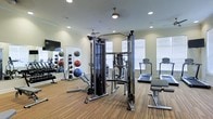 This fitness center offers a thoughtfully designed workout environment suited for a wide range of exercise routines. The space features warm wood-toned flooring that adds a welcoming feel, complemented by neutral walls and recessed lighting that ensure bright, even illumination throughout. A ceiling fan promotes comfortable airflow during workouts.

The equipment selection is extensive and well-organized. A multi-functional cable machine serves as the centerpiece, ideal for strength training and functional movement exercises. Alongside it, a selection of cardio machines — including treadmills and ellipticals — lines the perimeter near large windows that allow natural light to filter in. A dedicated free weight area features a full rack of dumbbells, while a medicine ball rack stocked with various weighted balls supports functional and core training.

Large wall-mounted mirrors enhance the sense of space and allow for proper form monitoring during exercise. A flat-screen television is mounted on the wall, providing entertainment during workouts. The single-floor layout ensures easy accessibility, and the thoughtful arrangement of equipment maximizes the use of floor space while maintaining open, comfortable movement areas. Overall, the fitness center provides a clean, functional, and well-lit environment for a complete workout experience.
