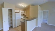 This single-floor residence offers a practical and comfortable layout designed for everyday living. The kitchen serves as a central feature of the home, equipped with white appliances including a full-size refrigerator and an electric range. Generous upper cabinetry with a warm wood trim provides ample storage, while dark countertops offer a subtle contrast to the light-colored walls and white cabinetry finishes. A peninsula-style breakfast bar with a matching dark countertop extends into the adjoining living area, creating a natural division between spaces while maintaining an open, connected feel. Track lighting above the kitchen provides focused illumination throughout the cooking area. The transition from the tiled kitchen floor to plush carpeting in the adjacent living space adds warmth and distinction between functional zones. Neutral, warm-toned walls throughout contribute to a welcoming ambiance. Additional doors visible from the main area suggest access to further rooms, such as bedrooms or storage, rounding out this well-organized single-level home.