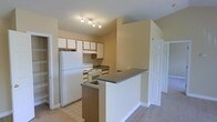 This single-floor residence offers a practical and comfortable layout designed for everyday living. The kitchen serves as a central feature of the home, equipped with white appliances including a full-size refrigerator and an electric range. Generous upper cabinetry with a warm wood trim provides ample storage, while dark countertops offer a subtle contrast to the light-colored walls and white cabinetry finishes. A peninsula-style breakfast bar with a matching dark countertop extends into the adjoining living area, creating a natural division between spaces while maintaining an open, connected feel. Track lighting above the kitchen provides focused illumination throughout the cooking area. The transition from the tiled kitchen floor to plush carpeting in the adjacent living space adds warmth and distinction between functional zones. Neutral, warm-toned walls throughout contribute to a welcoming ambiance. Additional doors visible from the main area suggest access to further rooms, such as bedrooms or storage, rounding out this well-organized single-level home.
