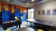 This dynamic, single-floor recreational space is designed with entertainment and social interaction in mind. The room makes an immediate visual impact with a striking, large-scale chess-themed mural spanning one full wall, rendered in vivid blue and orange tones that energize the entire space. The mural serves as a bold artistic centerpiece, complemented by framed chess-inspired artwork displayed on an adjacent neutral-toned wall.

The game room is well-equipped for active recreation, featuring a full-size ping-pong table positioned centrally to allow ample playing space on all sides, as well as a foosball table that adds to the variety of gaming options available. A high-top table with modern yellow accent chairs provides a casual seating area, ideal for spectators or between-game socializing.

Natural light filters into the space through a large window overlooking a lush outdoor tree line, balancing the room's lively interior palette with a calming exterior view. The flooring consists of a patterned carpet in muted tones, which grounds the space and provides comfort underfoot. Recessed ceiling lighting, paired with a distinctive curved teal ceiling detail, adds architectural interest overhead.

Overall, the space combines playful design elements with functional recreational amenities, creating an engaging and welcoming environment suited for leisure and community activity.