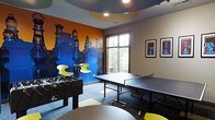 This dynamic, single-floor recreational space is designed with entertainment and social interaction in mind. The room makes an immediate visual impact with a striking, large-scale chess-themed mural spanning one full wall, rendered in vivid blue and orange tones that energize the entire space. The mural serves as a bold artistic centerpiece, complemented by framed chess-inspired artwork displayed on an adjacent neutral-toned wall.

The game room is well-equipped for active recreation, featuring a full-size ping-pong table positioned centrally to allow ample playing space on all sides, as well as a foosball table that adds to the variety of gaming options available. A high-top table with modern yellow accent chairs provides a casual seating area, ideal for spectators or between-game socializing.

Natural light filters into the space through a large window overlooking a lush outdoor tree line, balancing the room's lively interior palette with a calming exterior view. The flooring consists of a patterned carpet in muted tones, which grounds the space and provides comfort underfoot. Recessed ceiling lighting, paired with a distinctive curved teal ceiling detail, adds architectural interest overhead.

Overall, the space combines playful design elements with functional recreational amenities, creating an engaging and welcoming environment suited for leisure and community activity.
