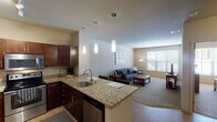 This single-floor residence offers a thoughtfully designed open-concept layout that seamlessly connects the kitchen and living areas. The kitchen is a standout feature, equipped with rich dark-wood cabinetry, granite countertops, stainless steel appliances including a refrigerator, range, and microwave, and a peninsula island with bar seating for two. Pendant lighting suspended from the ceiling adds a warm, focused glow over the workspace, while track lighting provides additional illumination throughout the kitchen. The adjacent living area is bright and inviting, anchored by a large window that allows ample natural light to fill the carpeted space. Two bedrooms provide comfortable accommodations, each featuring large windows that invite natural light and closet storage — one with sliding mirror-panel doors revealing a built-in rod and shelving system. The bathroom is clean and functional, featuring a combination tub and shower, a vanity with storage, and wood-look flooring. Neutral warm tones on the walls and light-colored ceilings throughout the residence create a cohesive, welcoming ambiance. The open floor plan and consistent finishes give this space a polished, move-in-ready feel.