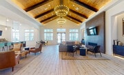 This thoughtfully designed clubhouse blends rustic warmth with modern sophistication across a single floor. The centerpiece is a grand main lounge with soaring vaulted ceilings clad in warm wood planks and accented by dark exposed beams. Dual statement chandeliers hang overhead, casting a welcoming glow over light wood-style flooring. A full-height brick fireplace wall anchors the seating area, which is furnished with mid-century modern chairs and plush sofas arranged on a vintage-style area rug. Banks of large windows and transom lights flood the space with natural light and offer outdoor views.

Adjacent to the lounge is a more intimate gathering area with a kitchenette, featuring a dark-topped counter bar with seating, stainless steel appliances, and shiplap wall detailing. Pendant lighting and high ceilings continue the cohesive design language.

A dedicated conference room provides a professional setting with a large wood table, ergonomic seating, a wall-mounted display, built-in cabinetry with storage, and glass partition doors that look into the fitness center beyond. The workspace area includes a built-in desk with a computer workstation.

A bright dining nook with whitewashed flooring, large windows, and a round table completes the layout. Throughout, shiplap walls, dark structural accents, and carefully curated artwork create a refined yet approachable atmosphere.