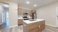 This well-designed single-floor unit offers a clean, contemporary aesthetic throughout its open and functional layout. The heart of the space is the modern kitchen, which features warm wood-toned cabinetry, crisp white countertops, and a full suite of stainless steel appliances including a gas range, over-the-range microwave, and refrigerator. A central kitchen island with an integrated sink adds both workspace and visual appeal, anchoring the open-concept living area. Light wood-style luxury vinyl plank flooring flows seamlessly through the main living and kitchen areas, creating a bright, cohesive feel.

The living area is generously proportioned, offering an open and airy atmosphere with recessed lighting and neutral warm-toned walls throughout. The bedrooms are finished with plush wall-to-wall carpeting and feature individual windows that allow for natural light and views of the surrounding area, along with dedicated climate control units.

Two bathrooms serve the space, each finished with large-format tile flooring and wood-toned vanities topped with white countertops. One bathroom includes a walk-in glass-enclosed shower, while the other provides additional utility. Together, the spaces present a cohesive, move-in ready environment with a contemporary design sensibility.