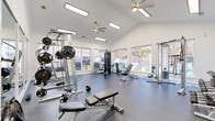 Located at 2600 Chestnut Oak Way in Columbus, Ohio, this single-floor fitness center offers a comprehensive workout facility thoughtfully divided into distinct training zones. The space is characterized by soaring vaulted ceilings, crisp white walls, and an abundance of natural light streaming through large, multi-pane windows that frame views of the surrounding landscape.

The main strength training area is spacious and open, housing a multi-station cable machine, adjustable weight benches, a barbell rack with free weights, a dumbbell rack, and a cable/pulley tower. Ceiling fans overhead ensure comfortable airflow throughout workouts. Durable rubber flooring covers the entire fitness floor, providing safety and sound absorption.

A dedicated cardio section features treadmills, elliptical trainers, and a stationary bike, all positioned beneath elegant arched windows with a wall-mounted flat-screen TV for entertainment. Full-length mirrors line one wall, enhancing the sense of space and allowing users to monitor their form.

A separate aerobics room, clearly labeled with frosted glass doors, provides a private area for group or individual aerobic exercise. The overall ambiance is bright, clean, and functional, offering a professional fitness environment well-suited to a variety of training needs.