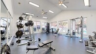 Located at 2600 Chestnut Oak Way in Columbus, Ohio, this single-floor fitness center offers a comprehensive workout facility thoughtfully divided into distinct training zones. The space is characterized by soaring vaulted ceilings, crisp white walls, and an abundance of natural light streaming through large, multi-pane windows that frame views of the surrounding landscape.

The main strength training area is spacious and open, housing a multi-station cable machine, adjustable weight benches, a barbell rack with free weights, a dumbbell rack, and a cable/pulley tower. Ceiling fans overhead ensure comfortable airflow throughout workouts. Durable rubber flooring covers the entire fitness floor, providing safety and sound absorption.

A dedicated cardio section features treadmills, elliptical trainers, and a stationary bike, all positioned beneath elegant arched windows with a wall-mounted flat-screen TV for entertainment. Full-length mirrors line one wall, enhancing the sense of space and allowing users to monitor their form.

A separate aerobics room, clearly labeled with frosted glass doors, provides a private area for group or individual aerobic exercise. The overall ambiance is bright, clean, and functional, offering a professional fitness environment well-suited to a variety of training needs.