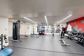 Located at 8750 Georgia Ave in Silver Spring, MD, this fitness center offers a generously sized, single-floor exercise space designed for a comprehensive workout experience. The facility is defined by a striking wall of floor-to-ceiling windows framed in red-accented mullions, flooding the interior with abundant natural light and providing open views of the surrounding urban greenery.

The cardio zone is well-equipped with a row of modern treadmills, elliptical trainers, and stationary bikes, all positioned along the expansive window wall to make the most of the outdoor views during workouts. The strength training area features a full rack of dumbbells, adjustable benches, a cable and weight machine system, and a dedicated free weights section complete with barbells and plate-loaded equipment. Full-length mirrors amplify the sense of space throughout the strength zone.

The interior design is contemporary and energetic, highlighted by a distinctive hexagonal acoustic tile feature wall in shades of red, gray, and white that adds a bold visual identity to the space. Durable rubber flooring runs throughout, providing safety and comfort underfoot. Overhead, the high ceilings and recessed lighting contribute to an open, airy atmosphere. A reception or check-in nook with vibrant green wall accents completes the facility's welcoming layout.
