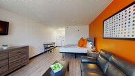 This Extra Large Suite in Memphis, Tennessee offers a generously proportioned, open-plan layout designed for both comfort and functionality. The suite's most striking design element is a bold, vibrant orange accent wall that anchors the sleeping and lounge area, creating a warm and energetic atmosphere throughout the space. Light wood-style flooring runs throughout, complementing the clean white walls and textured ceiling.

The sleeping area features a well-appointed bed with coordinating orange accent pillows, a rustic wood-framed headboard, and a matching nightstand with built-in lighting. Adjacent to the bed, a charcoal leather loveseat and a coffee table provide a comfortable lounge area, ideal for relaxing. A large wall-mounted flat-screen television sits above a six-drawer wooden dresser, offering ample storage. A compact dining table with chairs is conveniently positioned near the kitchenette area.

The kitchenette is outfitted with white cabinetry, a stainless steel sink, an over-the-counter microwave, a full-size refrigerator, and generous counter space — providing practical self-catering capabilities. A dedicated open closet area adds additional storage convenience.

The bathroom features a wide vanity with a stone-finish countertop, a large mirror, and a shower with a curtain accented in orange, tying cohesively into the suite's overall color scheme.