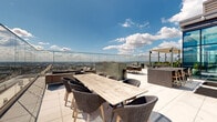 This expansive rooftop terrace offers a sophisticated outdoor experience elevated high above the surrounding cityscape. Enclosed by seamless glass railings that preserve unobstructed panoramic views, the terrace showcases sweeping vistas of the city skyline, a broad river, and open sky in every direction — a defining feature of this remarkable space.

The terrace is organized into several distinct functional zones across its generous layout. A central dining area features a large, rustic reclaimed wood table paired with woven rattan chairs, ideal for group gatherings. Adjacent to this, a comfortable lounge area is furnished with upholstered sofas and accent tables, providing a relaxed setting to take in the views. A dedicated bar zone with a pergola-style canopy and high stools offers an additional social focal point.

An outdoor kitchen station, equipped with a stainless steel gas grill and built-in cabinetry, makes the space well-suited for entertaining and al fresco dining. Integrated planter boxes filled with greenery serve as natural dividers between zones, adding organic texture to the sleek, contemporary design. 

Warm wood-toned steps with glass and steel railings connect tiered sections of the terrace, where sun loungers invite relaxation. The floor-to-ceiling glazed façade of the building anchors the space architecturally, reflecting the sky and reinforcing the terrace's elevated, urban character throughout.