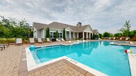 Located at 2038 SC-60 in Columbia, South Carolina, this two-story residential community is designed with a craftsman-inspired aesthetic, combining board-and-batten siding, stone accents, and a pitched shingle roof that gives the property a refined, welcoming character. The centerpiece of the outdoor amenity space is a generously sized swimming pool with crystal-clear water, ranging from shallow to mid-depth zones, and finished with clean white coping and a paver surround. Adjacent to the main pool is a separate wading or tanning ledge area, ideal for lounging in the water. White chaise lounge chairs and seating are arranged throughout the expansive pool deck, offering ample space for relaxation. The clubhouse building anchors one end of the pool area and features a covered rear porch with ceiling fans, stone fireplace, and outdoor seating — creating a comfortable gathering space in all seasons. Manicured Italian cypress trees frame the building and contribute to the polished landscaping. A second pool area with teal umbrella-shaded seating and additional lounge chairs provides further resort-style amenities. The surrounding multi-story residential buildings feature balconies and a complementary architectural palette of siding and stone, creating a cohesive community environment throughout.
