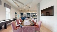 This well-appointed single-floor amenity space is designed to accommodate both social gatherings and focused work, offering a diverse range of functional zones within a cohesive, contemporary aesthetic. The main lounge area features soaring ceilings with a striking tray detail finished in deep blue, illuminated by a sculptural sputnik chandelier. Bold red and navy upholstered seating is arranged around a central coffee table, with large floor-to-ceiling windows and glass doors providing generous natural light and outdoor views of a pool area. Light wood-look flooring runs throughout, unifying the space.

The game room is defined by an elegant coffered ceiling and hosts a dedicated poker table surrounded by blush-pink upholstered chairs alongside a shuffleboard table, making it a standout entertainment hub. Adjacent to the game area, a full-length bar island with a marble-look countertop, woven barstools, and a textured blue tile backsplash serves as a social focal point, complete with pendant lighting and a wall-mounted television.

A private conference room offers a more intimate setting, featuring textured blue-grey wallcovering, a wall-mounted display, and a compact table with seating for small meetings or focused work. Throughout, curated artwork and considered color palettes elevate the overall ambiance.