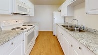 This single-floor residence offers a thoughtfully laid-out interior with a blend of hard-surface and carpeted flooring throughout. The spacious living area is a standout feature, showcasing warm hardwood-style flooring, dual ceiling fans, and a large wall-to-wall mirror that amplifies the sense of space and reflects abundant natural light. Sliding glass doors open to an outdoor area, allowing greenery views and excellent ventilation. Adjacent to the living space, the galley-style kitchen is efficiently designed with white cabinetry, granite-look countertops, a double stainless steel sink, and white appliances including a range with overhead microwave and a full-size refrigerator. The hardwood-style flooring continues seamlessly into the kitchen. The two bedrooms are comfortable and well-lit, each featuring plush carpeting, ceiling fans with light fixtures, and ample wall space. One bedroom benefits from a window with a view of surrounding foliage. The bathroom is clean and functional, appointed with mosaic tile flooring, white tile surround, a soaking tub, and a vanity with a granite-look countertop and white cabinetry. The overall ambiance of the space is bright, neutral, and move-in ready.