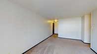This single-floor space offers a clean and functional interior layout designed for practical everyday use. The main living area is finished with light neutral carpeting that extends throughout, providing a soft and comfortable underfoot feel. The walls feature white vertical paneling that lends a simple, uniform aesthetic to the interior, while dark baseboards provide a subtle contrast along the perimeter. The textured white ceiling adds a classic touch and helps diffuse the ambient light throughout the room. A flush-mount ceiling fixture provides overhead lighting, contributing to the warm, even illumination of the space. Toward the rear of the living area, the flooring transitions to hardwood or laminate planking near the entryway, which is anchored by a solid dark-toned door with standard hardware. This transition zone between the carpeted living space and the hard-surface entry creates a natural delineation within the floor plan. The overall ambiance is straightforward and understated, offering a blank-canvas quality that lends itself well to personalization. The open, uncluttered layout makes the space feel airy and adaptable for a variety of uses.
