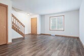 Located at 6801 Chester Dr in Madison, Wisconsin, this two-bedroom, one-bathroom home spans three floors with a practical and well-organized layout. The ground level houses an attached garage with a paneled overhead door and a utility room, providing convenient storage and vehicle access. The main living level features an open-concept living room and kitchen connected by a breakfast bar counter. The living room showcases durable hardwood-style flooring, warm wood baseboards, and a ceiling fan, while a carpeted staircase with a wood banister leads to the upper floor. The kitchen is outfitted with rich dark wood cabinetry, granite-style countertops, stainless steel appliances including a range and refrigerator, and hardwood-style flooring. The upper floor hosts two bedrooms, a hallway, two closets, and a full bathroom. The generously sized primary bedroom features plush carpet and a large walk-in closet equipped with multi-tier wire shelving. The second bedroom offers hardwood-style flooring and a wide window with outdoor views. The bathroom includes a white vanity with a large mirror and overhead lighting. Neutral tones, warm wood accents, and natural light from multiple windows contribute to the home's clean, functional ambiance throughout.