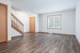 Located at 6801 Chester Dr in Madison, Wisconsin, this two-bedroom, one-bathroom home spans three floors with a practical and well-organized layout. The ground level houses an attached garage with a paneled overhead door and a utility room, providing convenient storage and vehicle access. The main living level features an open-concept living room and kitchen connected by a breakfast bar counter. The living room showcases durable hardwood-style flooring, warm wood baseboards, and a ceiling fan, while a carpeted staircase with a wood banister leads to the upper floor. The kitchen is outfitted with rich dark wood cabinetry, granite-style countertops, stainless steel appliances including a range and refrigerator, and hardwood-style flooring. The upper floor hosts two bedrooms, a hallway, two closets, and a full bathroom. The generously sized primary bedroom features plush carpet and a large walk-in closet equipped with multi-tier wire shelving. The second bedroom offers hardwood-style flooring and a wide window with outdoor views. The bathroom includes a white vanity with a large mirror and overhead lighting. Neutral tones, warm wood accents, and natural light from multiple windows contribute to the home's clean, functional ambiance throughout.