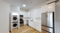 This single-floor unit presents a clean, modern aesthetic with warm hardwood-style flooring and crisp white walls carried consistently throughout. The kitchen serves as a functional centerpiece, outfitted with white shaker-style cabinetry featuring matte black hardware, white countertops, and a full suite of stainless steel appliances, including a French-door refrigerator, electric range, built-in microwave, and dishwasher. A subway tile backsplash adds texture behind the sink area, and a stacked washer and dryer unit is conveniently integrated into the kitchen space. The living area is open and bright, benefiting from natural light through a window overlooking an exterior balcony, and is equipped with a ceiling fan. The bedroom mirrors this brightness, featuring its own ceiling fan, a window with blinds, and a closet. The bathroom offers a tub-shower combination accented by a bold geometric patterned tile in black and white, providing a striking visual contrast to the otherwise minimal, white surroundings. The overall ambiance is fresh, functional, and contemporary.
