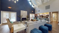 Located at 1700 Charleston Place Ln in Charlotte, NC, this single-story community clubhouse offers a thoughtfully designed gathering space for residents. The centerpiece is a grand open-concept great room distinguished by soaring vaulted ceilings with exposed beams, an elegant chandelier, and abundant natural light streaming through multiple French doors. Bold navy blue accent walls contrast with crisp white ceilings, while rich hardwood-style flooring runs throughout. A floor-to-ceiling stone fireplace serves as a dramatic focal point, flanked by stylish seating arrangements that include plush sofas, patterned armchairs, and cobalt blue ottomans. Adjacent to the main lounge is a full kitchen area outfitted with stainless steel appliances, a tiled bar counter with seating, and pendant lighting. A dedicated business center features built-in granite-topped workstations with computer terminals and a separate seating nook with a round table, all enclosed by glass partitions. A secondary lounge area provides a more relaxed setting with a wall-mounted television and mid-century style accent chairs. Throughout the space, French doors open directly to views of the outdoor pool, seamlessly connecting the interior with the surrounding outdoor amenities.
