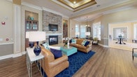 This single-floor community clubhouse is thoughtfully designed to offer residents a sophisticated and comfortable gathering space. The main lounge area is anchored by rich hardwood flooring, decorative columns with crown molding, and abundant natural light streaming through multiple white-framed windows. Plush seating arrangements in warm leather tones, accented with navy and teal throw pillows, are complemented by bold area rugs and elegant gold-framed coffee tables, creating an inviting and stylish atmosphere.

A standout feature of the space is the living room with a dramatic tray ceiling, a stacked-stone fireplace flanked by built-in display cabinets, and a mounted flat-screen television — a refined focal point that adds warmth and character. Adjacent seating areas with coordinating furnishings extend the social space seamlessly.

The kitchen is well-appointed with rich wood cabinetry, dark granite countertops, a stainless steel refrigerator, mosaic tile backsplash, and pendant lighting. A peninsula counter with bar stools offers a casual seating option, while a double-basin sink and ample counter space make it fully functional for events and gatherings.

A dedicated business center with workstations and seating provides a productive environment for residents. Throughout the space, consistent warm beige walls, hardwood and tile flooring, and recessed lighting tie every area together into a cohesive, polished whole.