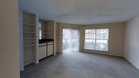 This single-floor space offers an open and airy layout designed for comfortable everyday living. The main living area is generously sized, featuring plush wall-to-wall carpeting that adds warmth underfoot. A standout feature of the space is the built-in shelving unit that divides the living area from the adjacent kitchen zone, providing both functional storage and a sense of architectural definition. The shelves offer ample display and organization options, while a dark countertop surface anchors the transitional space between the two areas. Natural light floods the interior through a large sliding glass door fitted with vertical blinds, which opens to an outdoor area and frames views of surrounding greenery. A wide picture window beside it further enhances the brightness of the room, creating a cheerful and welcoming ambiance throughout the day. The textured popcorn ceiling adds a classic residential character to the space. Overall, the layout balances openness with practical zoning, making it a functional and well-lit environment suitable for a variety of living arrangements.