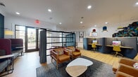 This thoughtfully designed co-working and lounge space offers a dynamic blend of collaborative and focused work environments across a single floor. The light-filled interior features warm hardwood flooring throughout, crisp white walls accented by a bold teal feature wall, and recessed lighting complemented by colorful pendant lamps in blue, yellow, and red tones.

The lounge area is anchored by a set of tan leather armchairs with colorful throw pillows arranged around a sculptural marble-top coffee table on a textured area rug. High-back booth-style seating upholstered in burgundy and grey fabric lines the teal accent wall, providing semi-private nooks adorned with decorative wall art and warm sconce lighting overhead.

Dedicated workstations are housed within curved, acoustically designed dark grey pod dividers, each equipped with a computer monitor and yellow task chairs, offering focused privacy within the open layout. Additional seating is available at a round table with cane-back chairs.

A sleek black steel shelving unit serves as a visual divider near glass-panel doors that open to an outdoor balcony, allowing natural light to pour in. Bold geometric black-and-white murals and a colorful hexagonal art installation add artistic character to the space, creating an inspiring and functional environment suited for both productivity and relaxation.
