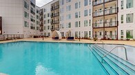 Located at 4101 Raleigh St in Charlotte, NC, this outdoor amenity space is centered around a generously sized resort-style swimming pool set within a landscaped courtyard. The pool is enclosed on multiple sides by a modern multi-story building featuring a crisp white façade with large windows, individual balconies, and bold pops of color — including a striking mural in terracotta, green, and cream tones on the parking structure backdrop. The pool deck is finished in warm-toned concrete and is well-appointed with a row of cushioned lounge chairs paired with striped umbrellas along one side, complemented by distinctive arched cabana-style daybeds that add a resort atmosphere. Lush landscaping with manicured shrubs and seasonal plantings lines the perimeter, softening the hardscape and adding greenery to the courtyard. An outdoor shower station and clearly posted pool rules signage are accessible near the pool entrance. Large glass French doors along the building's ground floor provide interior access to and from the courtyard, offering views of the pool from within. The space is designed to maximize open-air relaxation while maintaining an intimate, enclosed courtyard feel — ideal for recreation and leisure.