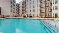 Located at 4101 Raleigh St in Charlotte, NC, this outdoor amenity space is centered around a generously sized resort-style swimming pool set within a landscaped courtyard. The pool is enclosed on multiple sides by a modern multi-story building featuring a crisp white façade with large windows, individual balconies, and bold pops of color — including a striking mural in terracotta, green, and cream tones on the parking structure backdrop. The pool deck is finished in warm-toned concrete and is well-appointed with a row of cushioned lounge chairs paired with striped umbrellas along one side, complemented by distinctive arched cabana-style daybeds that add a resort atmosphere. Lush landscaping with manicured shrubs and seasonal plantings lines the perimeter, softening the hardscape and adding greenery to the courtyard. An outdoor shower station and clearly posted pool rules signage are accessible near the pool entrance. Large glass French doors along the building's ground floor provide interior access to and from the courtyard, offering views of the pool from within. The space is designed to maximize open-air relaxation while maintaining an intimate, enclosed courtyard feel — ideal for recreation and leisure.
