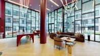 This contemporary lobby lounge makes an immediate impression with its dramatic double-height ceilings and expansive floor-to-ceiling glass curtain walls, flooding the interior with abundant natural light and offering open views of the surrounding courtyard and building exterior. The herringbone-patterned hardwood flooring runs throughout, lending warmth and visual texture to the generously proportioned space.

The lounge is thoughtfully organized into distinct seating zones, each with its own character. A central gathering area features a large sectional sofa in rich brown, paired with accent chairs and a wooden coffee table, anchored beneath a sculptural rope-style chandelier. Adjacent to this, a media area offers a wall-mounted large-screen television set against a dusty rose accent wall, complemented by additional comfortable seating.

A vibrant banquette area lines one wall with plush coral-toned bench seating and marble-top café tables, set against bold burgundy and teal color-blocked walls. Nearby, a striking red architectural table with arched legs serves as a social bar or gathering counter with barstool seating.

Bold crimson cylindrical columns punctuate the space, reinforcing the energetic, design-forward palette. The overall ambiance is modern, inviting, and visually dynamic — ideal for social interaction and community engagement.