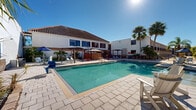 Located at 3020 Reedy Creek Blvd in Kissimmee, Florida, this outdoor pool area embodies the relaxed, sun-soaked lifestyle of Central Florida. The centerpiece of the space is a generously sized swimming pool with crystal-clear turquoise water, surrounded by a wide, paved pool deck finished in light-toned stone pavers that reflect the abundant Florida sunshine. Multiple pergolas and umbrella-shaded lounging areas are strategically positioned around the pool, offering guests comfortable spots to relax in the shade. Lounge chairs and seating are arranged throughout the deck, providing ample space for sunbathing and outdoor leisure. A decorative stone water feature wall adds a distinctive architectural element to the area, complementing the resort-style ambiance. Tall, swaying palm trees and lush tropical plantings frame the pool on all sides, enhancing the sense of a verdant, private oasis. The surrounding building features a Mediterranean-inspired design with a terracotta tile roof, white stucco exterior walls, and large windows, while upper-level balconies overlook the pool courtyard. The entire space is thoughtfully arranged to maximize outdoor enjoyment, making it an inviting and functional amenity well-suited to the warm Kissimmee climate.