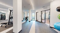This well-appointed fitness center offers a generous single-floor footprint designed to accommodate a full range of exercise activities. The space is divided into two primary zones, seamlessly connected by an open, airy layout that promotes ease of movement throughout.

The main workout area is defined by its clean, contemporary aesthetic — crisp white walls, structural columns, and continuous linear LED lighting that runs along the ceiling, providing consistent, bright illumination throughout. Durable, multi-toned carpet flooring with distinct striping delineates different functional areas and adds a professional finish underfoot.

The cardio zone is equipped with treadmills, elliptical machines, stationary bikes, and rowing equipment, positioned alongside large windows that offer natural light and outdoor views of lush greenery and palm trees. Full-length wall mirrors expand the sense of space and allow users to monitor their form.

The strength training zone features a comprehensive array of weight machines, squat racks, free-weight benches, and an organized rack of dumbbells, medicine balls, and resistance accessories. Built-in shelving with storage cabinetry keeps equipment neatly arranged.

A separate glass-walled room — visible from the main floor — provides a dedicated space for stretching or group fitness activities with hardwood flooring. The overall ambiance is modern, clean, and purposefully designed to support a premium fitness experience.