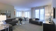 This single-floor space offers an open-concept layout that seamlessly integrates the living and dining areas into one cohesive, well-lit environment. Large windows along multiple walls flood the interior with abundant natural light, creating an airy and welcoming ambiance throughout. The living area is anchored by two charcoal-grey sofas arranged around a sleek circular glass coffee table, providing a comfortable and stylish setting for relaxation or entertaining. Soft, neutral-toned carpeting runs throughout the space, contributing to a warm and understated aesthetic. Track lighting overhead supplements the generous natural light, ensuring the interior remains well-illuminated at all times. The dining area, positioned adjacent to the living space, features a dark-toned table paired with upholstered dining chairs, offering a functional yet refined area for meals. White drapery panels frame the windows, adding an elegant finishing touch while allowing light to filter through. The overall design is contemporary and cohesive, characterized by a restrained neutral palette of grays and whites that lends the space a clean, modern feel.