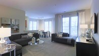 This single-floor space offers an open-concept layout that seamlessly integrates the living and dining areas into one cohesive, well-lit environment. Large windows along multiple walls flood the interior with abundant natural light, creating an airy and welcoming ambiance throughout. The living area is anchored by two charcoal-grey sofas arranged around a sleek circular glass coffee table, providing a comfortable and stylish setting for relaxation or entertaining. Soft, neutral-toned carpeting runs throughout the space, contributing to a warm and understated aesthetic. Track lighting overhead supplements the generous natural light, ensuring the interior remains well-illuminated at all times. The dining area, positioned adjacent to the living space, features a dark-toned table paired with upholstered dining chairs, offering a functional yet refined area for meals. White drapery panels frame the windows, adding an elegant finishing touch while allowing light to filter through. The overall design is contemporary and cohesive, characterized by a restrained neutral palette of grays and whites that lends the space a clean, modern feel.