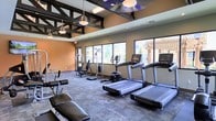 This fitness center is designed to provide a comprehensive workout experience within a thoughtfully appointed single-floor space. The room features striking exposed dark structural beams across the ceiling, adding an industrial aesthetic that contrasts with the warm terracotta-toned walls. Pendant lighting and recessed fixtures illuminate the space, complementing the natural light that pours in through a row of large windows along one wall. These windows also offer pleasant views of an outdoor pergola and surrounding greenery, creating an open, airy atmosphere during workouts. The floor is finished with durable, textured carpet tiles well-suited for a high-traffic fitness environment. The space is outfitted with a robust selection of modern exercise equipment, including multiple treadmills equipped with individual display screens, an upright stationary bike, a multi-function cable and weight machine, and a dedicated free weight and bench press station. The layout is spacious and well-organized, allowing users to move freely between stations without feeling crowded. Overall, this fitness center combines functional equipment with an inviting design, making it a practical and motivating environment for a wide range of fitness activities.