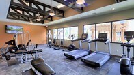 This fitness center is designed to provide a comprehensive workout experience within a thoughtfully appointed single-floor space. The room features striking exposed dark structural beams across the ceiling, adding an industrial aesthetic that contrasts with the warm terracotta-toned walls. Pendant lighting and recessed fixtures illuminate the space, complementing the natural light that pours in through a row of large windows along one wall. These windows also offer pleasant views of an outdoor pergola and surrounding greenery, creating an open, airy atmosphere during workouts. The floor is finished with durable, textured carpet tiles well-suited for a high-traffic fitness environment. The space is outfitted with a robust selection of modern exercise equipment, including multiple treadmills equipped with individual display screens, an upright stationary bike, a multi-function cable and weight machine, and a dedicated free weight and bench press station. The layout is spacious and well-organized, allowing users to move freely between stations without feeling crowded. Overall, this fitness center combines functional equipment with an inviting design, making it a practical and motivating environment for a wide range of fitness activities.