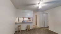 This single-floor residential space offers a practical and functional layout designed for comfortable everyday living. The living area is finished with wall-to-wall carpeting and features a ceiling fan with a light fixture, providing both airflow and ambient lighting. A textured ceiling adds a classic touch throughout the space.

The kitchen is efficiently designed with a pass-through counter that opens directly to the living area, creating a connected and sociable atmosphere. The counter is lined with wooden bar stools, making it ideal for casual dining or entertaining. The kitchen is equipped with essential appliances, including a refrigerator and a range, along with cabinetry for ample storage. A sink is positioned beneath the pass-through window, keeping the workspace practical and accessible.

A hallway visible from the living area leads to the additional rooms of the unit, which are accessible through paneled doors. The flooring transitions from carpet in the living area to tile in the connecting corridor, adding subtle variety to the overall aesthetic.

The space presents a clean, neutral palette throughout, with white walls and ceilings that lend a bright, open feel. The layout is straightforward and well-connected, making efficient use of the available floor plan across its single level.