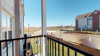 This single-floor residential unit offers a practical and well-organized layout designed for comfortable everyday living. The open-concept main living area seamlessly connects the living room and kitchen, creating a spacious, cohesive environment. The kitchen is equipped with rich, dark wood cabinetry, white appliances including a refrigerator and stove, and a peninsula island with a light wood finish that provides additional counter space and serves as a natural divider between the kitchen and living area. The flooring transitions from tile in the kitchen to plush carpeting in the living room, adding warmth underfoot. A dedicated entryway with a paneled door provides a clear sense of arrival. The carpeted bedroom is well-lit by large windows that invite abundant natural light and features a ceiling fan with a light fixture for added comfort. A sliding glass door opens directly onto a private balcony, which overlooks the surrounding neighborhood, open green lawns, and distant open land under a broad sky. The balcony itself is framed by black metal railings, offering a pleasant outdoor retreat. Throughout the unit, neutral wall tones and wood baseboards contribute to a clean, understated aesthetic that is easy to personalize.