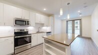 This efficiently designed 1-bedroom, 1-bathroom unit in Austin, Texas offers a thoughtfully laid-out floor plan that maximizes every square foot. The open-concept kitchen and living area serves as the heart of the home, highlighted by a central island with a warm-toned countertop and built-in shelving below. The kitchen is outfitted with stainless steel appliances—including a range, microwave, and dishwasher—complemented by crisp white cabinetry, subway tile backsplash, and light wood-style flooring that flows throughout the unit. Pendant lighting above the island adds a modern touch. The living area is bathed in natural light, with sliding glass doors opening to a private balcony offering views of the surrounding urban environment. The bedroom is a calm, neutral retreat featuring large windows that bring in ample daylight, a ceiling fan, and a dedicated closet. The bathroom is finished with large-format white floor tiles, a white vanity with warm brown countertop, a framed mirror, and a shower-tub combo with tile surround. Clean lines, a neutral color palette, and consistent wood-look flooring throughout create a cohesive and modern aesthetic well-suited to Austin living.