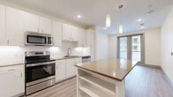 This efficiently designed 1-bedroom, 1-bathroom unit in Austin, Texas offers a thoughtfully laid-out floor plan that maximizes every square foot. The open-concept kitchen and living area serves as the heart of the home, highlighted by a central island with a warm-toned countertop and built-in shelving below. The kitchen is outfitted with stainless steel appliances—including a range, microwave, and dishwasher—complemented by crisp white cabinetry, subway tile backsplash, and light wood-style flooring that flows throughout the unit. Pendant lighting above the island adds a modern touch. The living area is bathed in natural light, with sliding glass doors opening to a private balcony offering views of the surrounding urban environment. The bedroom is a calm, neutral retreat featuring large windows that bring in ample daylight, a ceiling fan, and a dedicated closet. The bathroom is finished with large-format white floor tiles, a white vanity with warm brown countertop, a framed mirror, and a shower-tub combo with tile surround. Clean lines, a neutral color palette, and consistent wood-look flooring throughout create a cohesive and modern aesthetic well-suited to Austin living.
