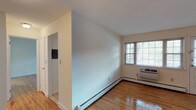 Located at Chelsea Village, 77, this single-floor residence offers a clean, functional layout designed for comfortable everyday living. Polished hardwood floors run throughout the main living areas and bedrooms, lending warmth and a classic character to the space. The living area is bright and open, benefiting from natural light streaming in through multiple windows fitted with blinds, and is equipped with baseboard heating and a wall-mounted air conditioning unit for year-round comfort. A front entry door provides direct access to the living space, adding convenience. The kitchen is well-appointed with rich, dark cherry wood cabinetry, granite-style dark countertops, a tile backsplash, and a full suite of black appliances including a gas range, microwave, and dishwasher. A window above the sink brings in additional natural light, making the kitchen a practical and pleasant workspace. The bedroom area continues the hardwood flooring and connects to additional rooms, providing flexible use of space. The bathroom features a tiled tub-and-shower combination enclosed by a sleek sliding glass door with chrome fixtures, complemented by neutral tile work for a clean, timeless look. Overall, the unit presents a well-maintained, move-in-ready space with consistent finishes and a straightforward, efficient floor plan.