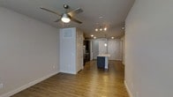This single-floor space showcases a thoughtfully designed open-concept layout that integrates the living and kitchen areas into one cohesive environment. Warm hardwood-style flooring runs throughout, lending a sense of continuity and warmth to the space.

The living area is equipped with a ceiling fan featuring an integrated light fixture, providing both comfort and ambient illumination. Neutral gray walls create a clean, contemporary backdrop that complements the natural tones of the flooring.

The kitchen stands out as a focal point, anchored by a central island with a built-in sink and navy blue cabinetry below, offering both functional workspace and a striking design contrast. Dark cabinetry lines the kitchen walls, housing stainless steel appliances, while pendant lighting above the island adds a refined, modern touch. Recessed lighting and a track lighting system ensure the kitchen remains well-lit throughout.

A sliding barn-style door and multiple interior doors provide access to additional areas of the space, while clean white trim and baseboards throughout reinforce the modern aesthetic. The layout promotes excellent sightlines and an open, airy atmosphere across the entire floor plan.