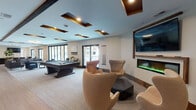 Located at 67–115 W Washington St in Oswego, Illinois, this single-floor community amenity space is designed for socializing and recreation. The layout is generously open, flowing seamlessly from one zone to the next across a combination of hardwood-style flooring and plush carpeting.

The main lounge area is anchored by a sleek electric fireplace mounted beneath a large wall-mounted television, creating a warm focal point. Surrounding it, a curated arrangement of sculptural egg-style lounge chairs and low-profile accent tables invites relaxed conversation. A dedicated game zone features a full-size billiards table and mounted cue rack, adding an energetic counterpoint to the lounge's relaxed atmosphere.

Throughout the space, a secondary seating area offers stylish dark upholstered sofas paired with glass-top coffee tables, accompanied by bar-height bistro tables and gold-accented stools. A kitchenette with dark cabinetry, light quartz countertops, and a double sink provides convenience for gatherings, with an additional wall-mounted TV nearby.

Architecturally, the space is distinguished by a coffered ceiling with dark wood inset panels and recessed lighting that adds depth and sophistication. Expansive floor-to-ceiling sliding glass doors line multiple walls, flooding the interior with natural light and offering views of the surrounding courtyard and streetscape.