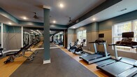Located at 401 Pearl St in Jeffersonville, Indiana, this well-appointed fitness center offers a comprehensive workout environment across a single, spacious floor. The facility is thoughtfully designed with a combination of hardwood and rubber flooring, clearly delineating the cardio and strength training zones.

The cardio section lines one side of the room and is equipped with multiple treadmills, stationary bikes, and elliptical machines, all positioned in front of large windows that invite natural light and offer outdoor views of the surrounding area. Ceiling fans are evenly distributed throughout, ensuring proper air circulation during workouts.

The free weights area features a full rack of dumbbells alongside adjustable benches, catering to a range of strength training exercises. Floor-to-ceiling mirrors span the walls in this section, creating a sense of expansiveness and allowing users to monitor their form. Structural columns with teal accents add a distinctive architectural character to the space.

On the opposite end of the room, a multi-station cable and weight machine provides options for full-body strength conditioning. The walls throughout the facility are finished in a two-tone palette of teal and warm gray, accented with geometric trim detailing that adds a modern, polished aesthetic. A glass-panel entry door with keypad access completes this well-equipped, professionally designed fitness center.