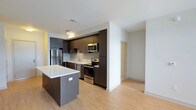 This single-floor space showcases a contemporary open-concept design centered around a well-appointed kitchen. Dark espresso cabinetry lines the walls from floor to ceiling, providing ample storage while creating a bold visual contrast against the white walls and light hardwood flooring that extends throughout the unit. The kitchen is fully equipped with stainless steel appliances, including a refrigerator, electric range, and over-the-range microwave, alongside a undermount sink set into light-colored countertops. A kitchen island with a white quartz surface and matching dark cabinetry serves as a functional centerpiece, offering additional workspace and casual seating potential. Recessed track lighting illuminates the kitchen area, while white walls and light flooring enhance the sense of openness throughout. A hallway visible beyond the main living area leads to additional rooms, suggesting a practical and efficient floor plan. The clean lines, neutral palette, and modern finishes combine to create a cohesive, move-in-ready living environment with a polished, urban aesthetic.