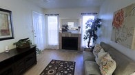 This single-floor residence offers a warm and functional living environment with a well-considered layout. The living room serves as the centerpiece of the home, featuring light hardwood-style flooring that extends throughout the space and a bold, tiled fireplace as the room's focal point. The fireplace surround is clad in earth-toned tile, adding both texture and visual interest to the area. Large windows dressed with sheer curtains flank the fireplace and the entryway, allowing generous natural light to fill the room and create an open, airy atmosphere. A decorative area rug with a bold circular pattern anchors the seating area, complementing the neutral wall tones and textured ceiling. Lush indoor plants are thoughtfully placed throughout, adding a touch of greenery and life to the interior. A pass-through view into the kitchen hints at a connected, open-concept design, where white cabinetry and bright lighting are visible in the background. The front entry features a classic paneled white door, contributing to the home's clean and welcoming aesthetic. Overall, the space presents a cozy yet bright environment with a cohesive design that balances comfort and practicality.