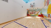 Located at 1515 Surf Ave in Brooklyn's Coney Island neighborhood, this indoor sports court is a dynamic and visually striking amenity space. The court features polished hardwood flooring marked with blue and yellow court lines, suitable for basketball and other indoor sports. A regulation basketball hoop with a clear backboard is mounted on the wall, maximizing the playability of the space.

The most distinctive architectural feature is the soaring ceiling height, which creates an open, airy atmosphere and allows for unrestricted athletic activity. A skylight element near the ceiling brings in natural light, further brightening the interior.

Dominating one end of the court is a bold, floor-to-ceiling mural celebrating the spirit of Coney Island. The artwork depicts iconic local imagery including a Ferris wheel, roller coasters, mermaids, and festive characters, all rendered in vivid, energetic colors. The "1515 Surf" address is prominently featured in the mural, tying the art directly to the building's identity.

A contrasting exposed concrete wall runs along one side, adding an industrial design element that balances the colorful mural. Stainless steel railings near the entry area provide safe access to the court level. Recessed and track lighting supplements the natural light, ensuring the space is well-illuminated for active use at all times.