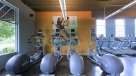 Located at 5704 New Albany Rd in West New Albany, Ohio, this two-story fitness facility offers a well-equipped and energizing environment designed to support a full range of workout routines. The space features an open floor plan with high ceilings and exposed industrial-style ductwork overhead, creating a spacious, gym-like atmosphere. Natural light filters in through tall windows, brightening the workout areas and contributing to an uplifting ambiance.

The cardio floor is outfitted with a robust selection of Precor ellipticals and treadmills, arranged in rows to maximize capacity and ease of movement between machines. Each machine is equipped with individual display screens, allowing users to track their workout metrics. Bold, large-scale athletic murals adorn the warm orange accent walls, adding visual motivation and energy to the space.

The flooring throughout features dark hardwood-style material, providing both durability and a polished aesthetic. A staircase connects the two levels, suggesting additional workout or functional spaces on the upper floor. The overall design balances industrial architecture with motivating visual elements, making this facility both functional and visually engaging for fitness enthusiasts.