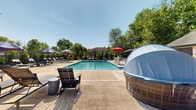 Located at 2101 Glen Dr in Cedar Knolls, New Jersey, this outdoor recreational amenity complex centers around a generously sized swimming pool with crystal-clear blue water. The pool ranges from a shallow end of 4 feet, making it accessible for a variety of swimmers, and is surrounded by a broad concrete pool deck that provides ample space for lounging and socializing.

The sundeck is well-appointed with a large collection of chaise lounge chairs and stylish dark-framed seating arranged along the pool's perimeter, all shaded by a row of bold purple umbrellas. Additional seating areas are scattered throughout the deck, including cozy pod-style lounge chairs, outdoor dining tables with umbrella coverage, and upholstered patio furniture, offering multiple settings for relaxation and gathering.

A charming single-story clubhouse building anchors the space, featuring a steeply pitched brown shingled roof, a cupola, and large windows that open toward the pool area. An attached covered patio with string lights extends the clubhouse's entertainment space outdoors. The complex is enclosed by a classic black iron fence and framed by lush mature trees, providing both privacy and a verdant backdrop. Manicured shrubs and potted plantings add greenery throughout the deck, enhancing the resort-like ambiance of this community amenity.